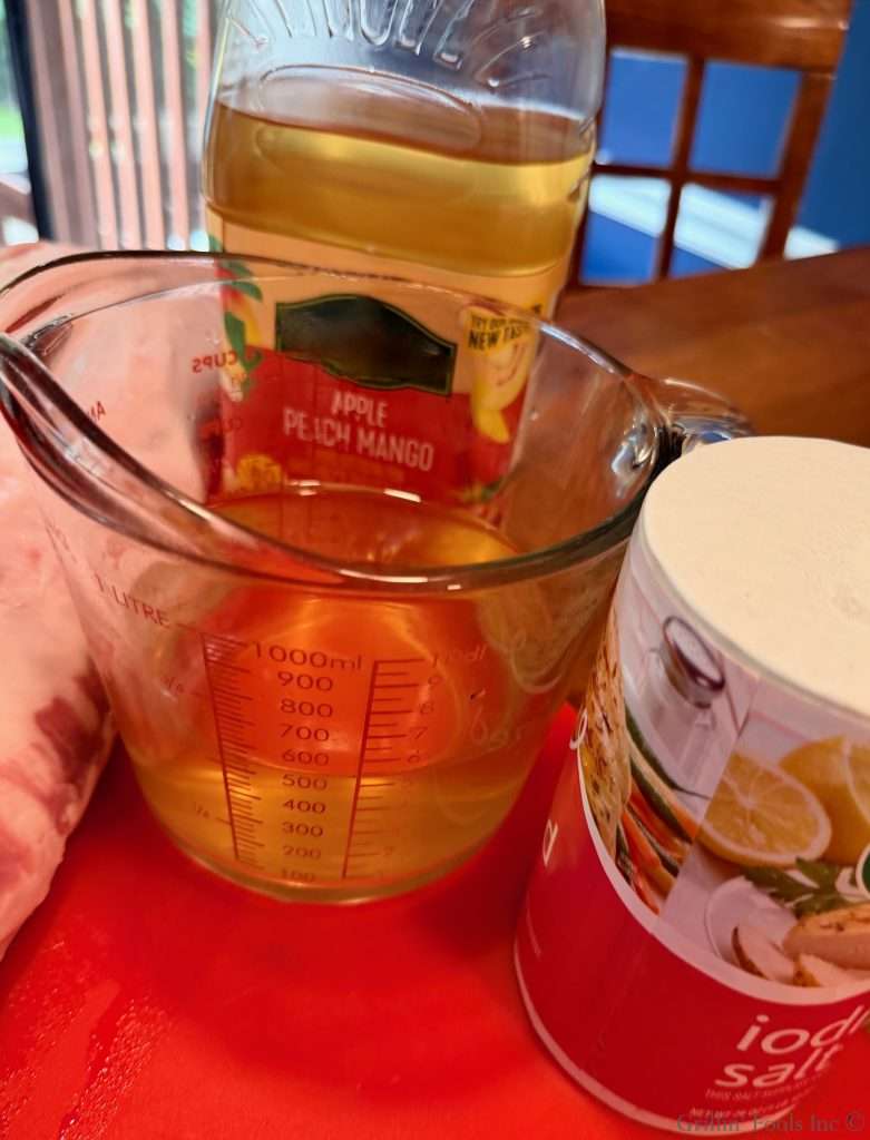 Measuring cup filled with a pale yellow liquid placed on a red surface, with a juice bottle labeled apple peach mango in the background and a container on the right.