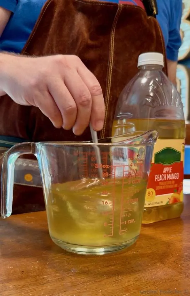Person in a brown apron stirs a pale yellow liquid in a glass measuring cup; a bottle labelled 'Apple Peach Mango' sits nearby on the counter.