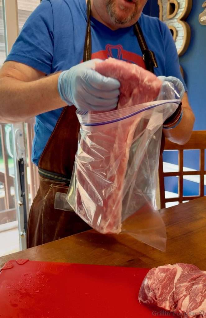 Butcher wearing gloves seals a large portion of raw meat in a plastic bag, with more meat on a red cutting board nearby.