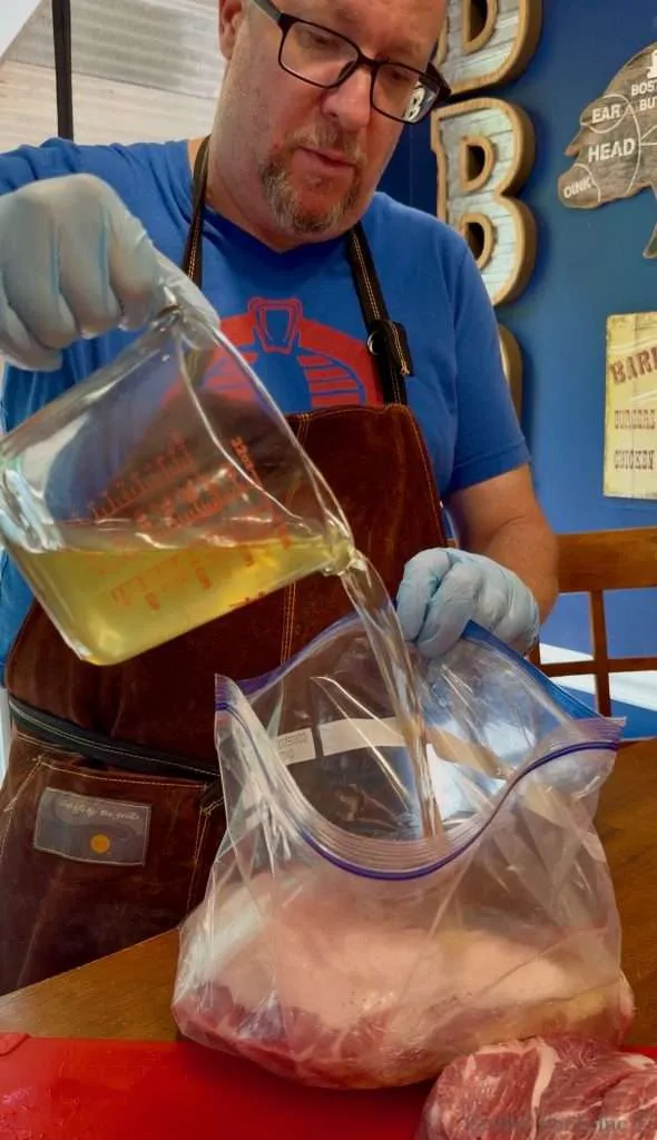 Butcher wearing an apron and gloves pours brine from a measuring cup into a large zip-top bag of meat on a wooden table.