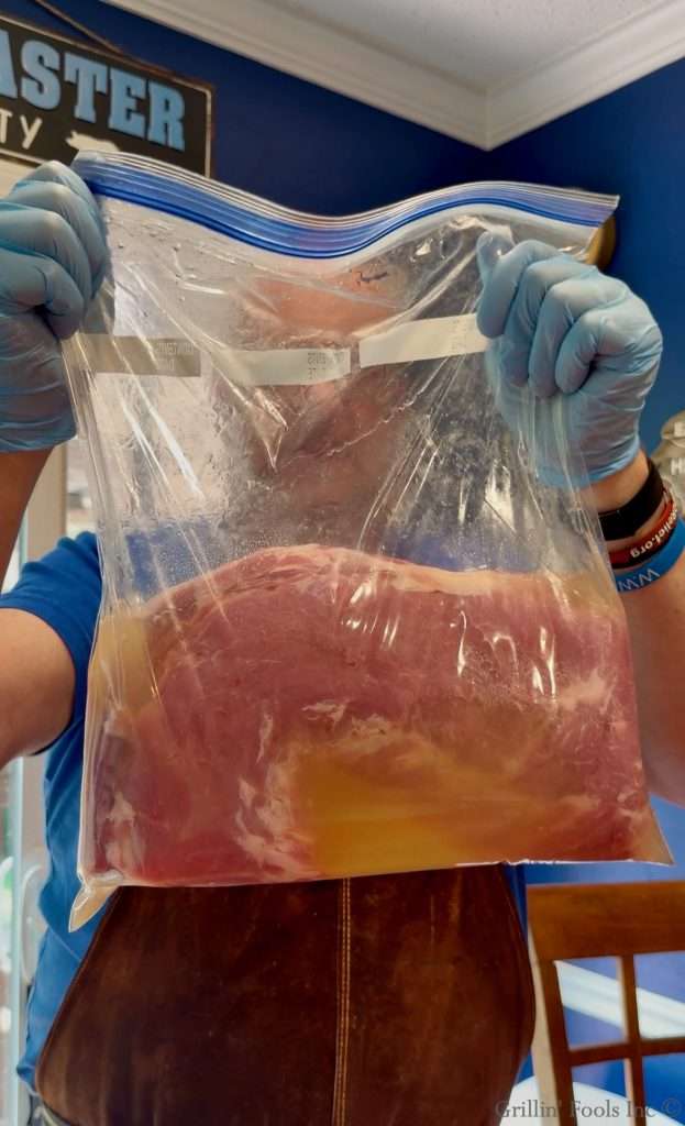 Person wearing blue gloves holds a large resealable bag containing pink meat soaked in yellow liquid for brining/marinating.