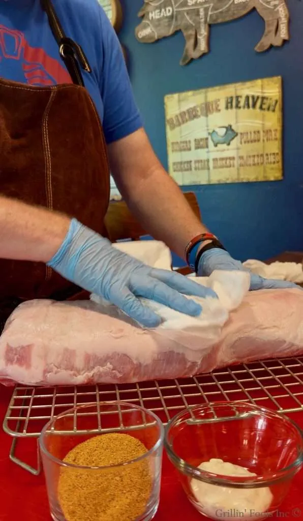 Butcher in blue shirt and apron trims a large raw pork loin on a rack, with bowls of spices nearby.