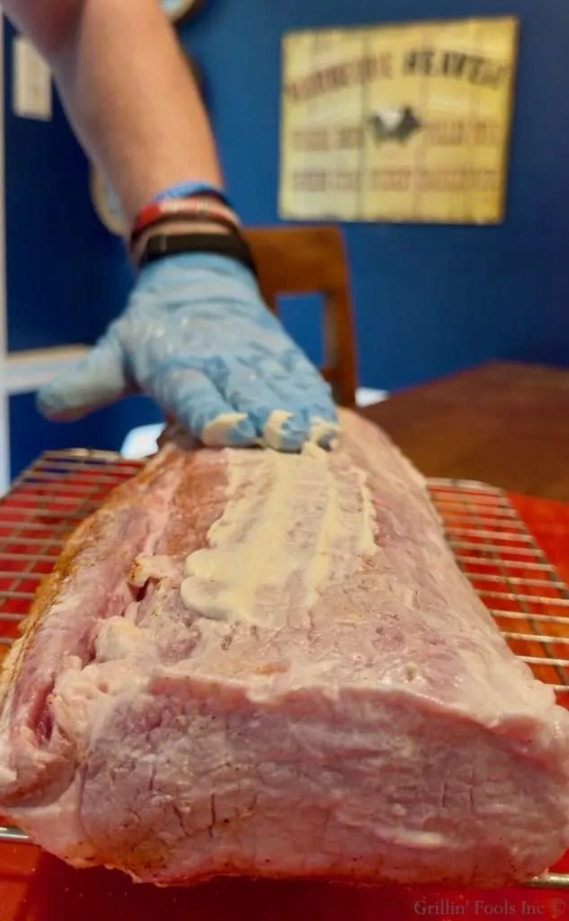 Gloved hand smoothing a layer of mayonnaise on a raw pork loin resting on a rack.