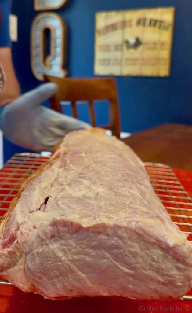 Raw pork roast, slathered with mayonnaise, resting on a wire rack in a kitchen, with a blue wall in the background