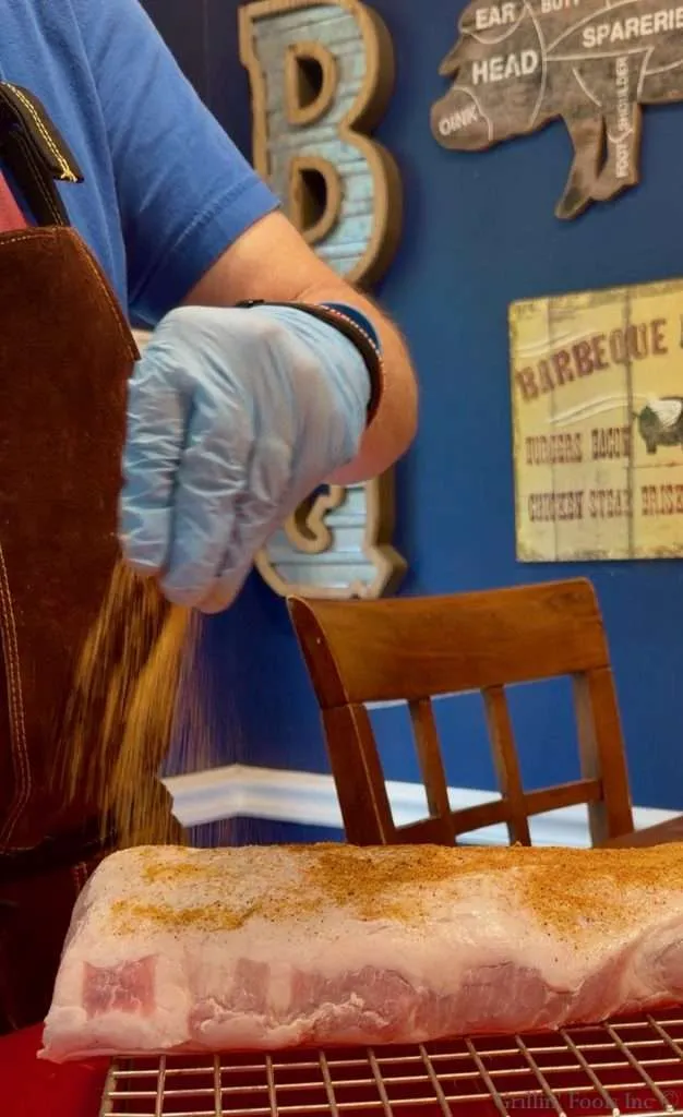 Chef wearing blue gloves sprinkles dry rub onto a slab of pork loin on a cookie cooling rack.
