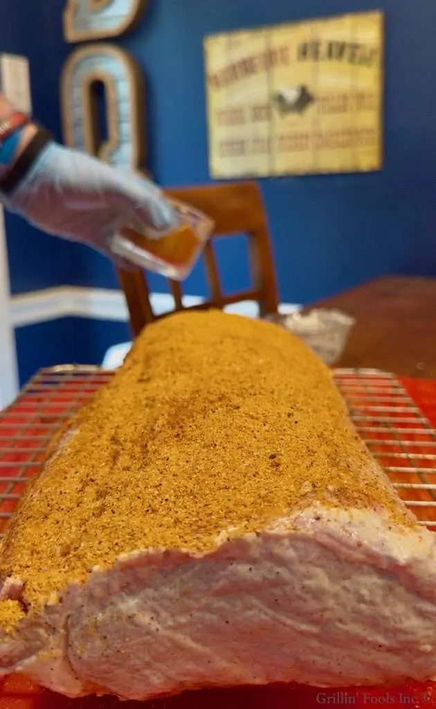 Close-up of a large seasoned pork roast on a rack, a gloved hand pouring sauce over it indoors.