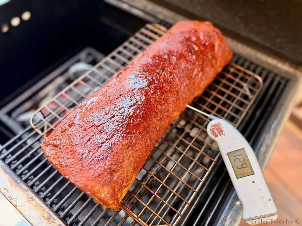 Barbecued pork roast on a grill grate with a digital thermometer showing 129°F.