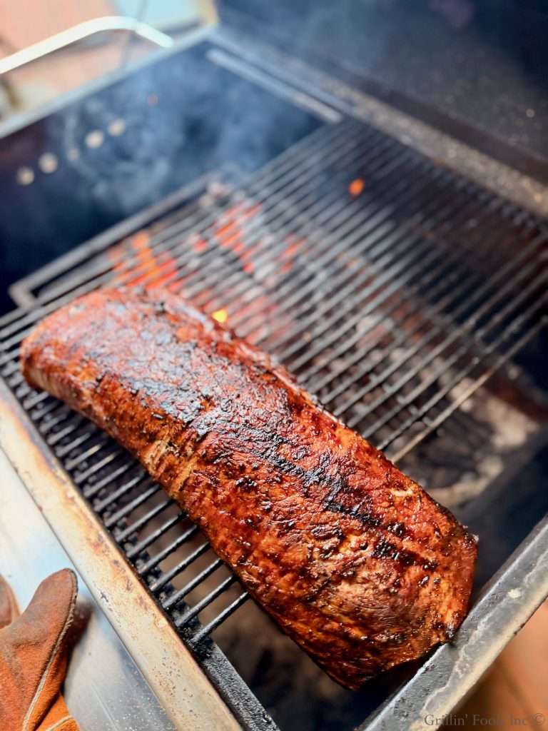 A large piece of barbecued pork with a dark, glazed bark cooking on a charcoal grill with flames and smoke rising nearby.