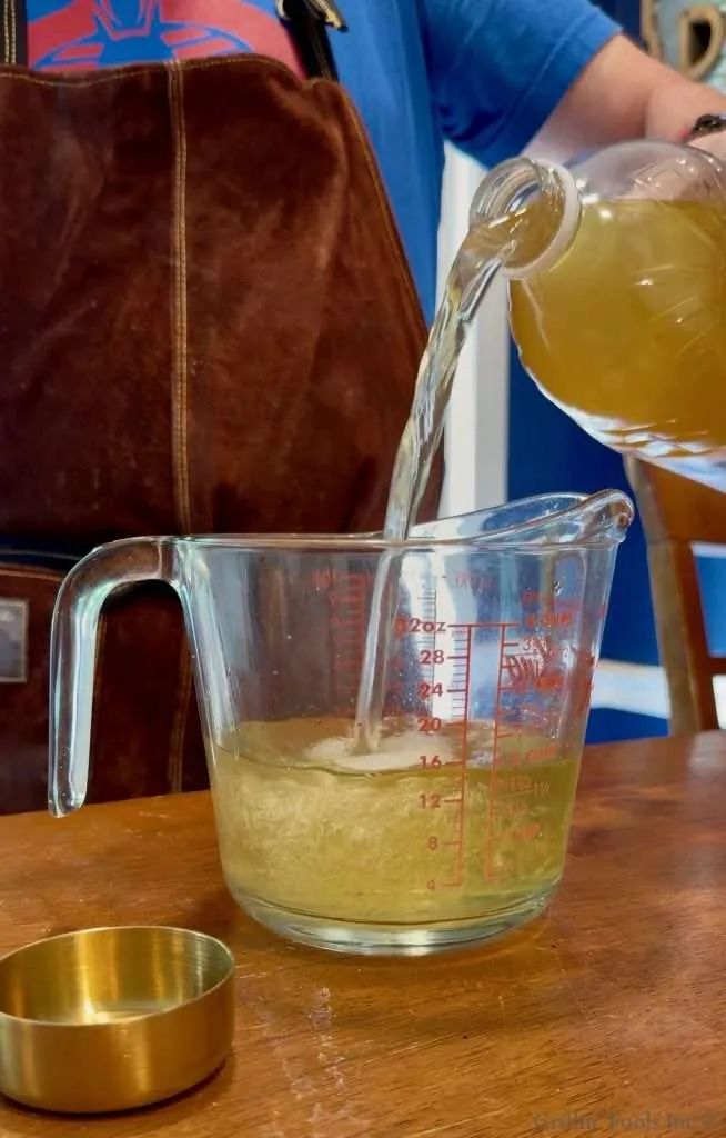 Person pours a yellow liquid from a pitcher into a clear measuring cup on a wooden table, wearing a blue shirt and brown apron