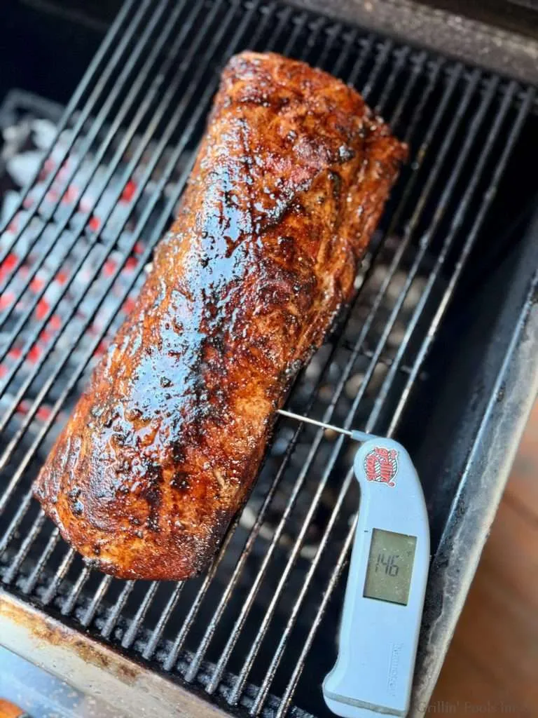 Barbecued pork loin cooking on grill with a digital meat thermometer reading 146°F.