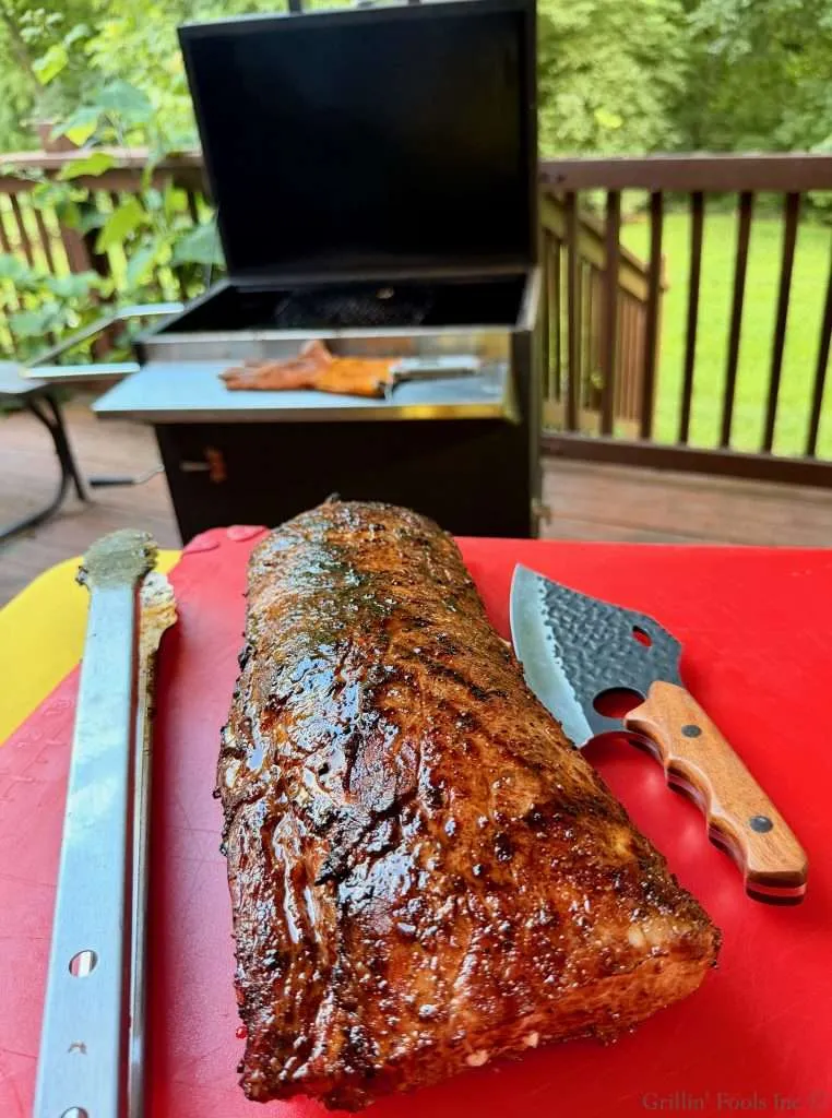 Smoked pork loin on a red cutting board with a knife, outdoors near a grill and green backyard.