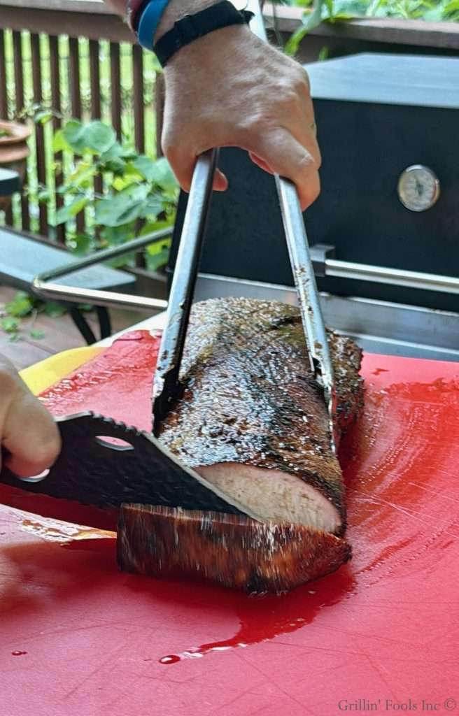 Hand using tongs to carve a thick smoked pork loin on a red cutting board outdoors.