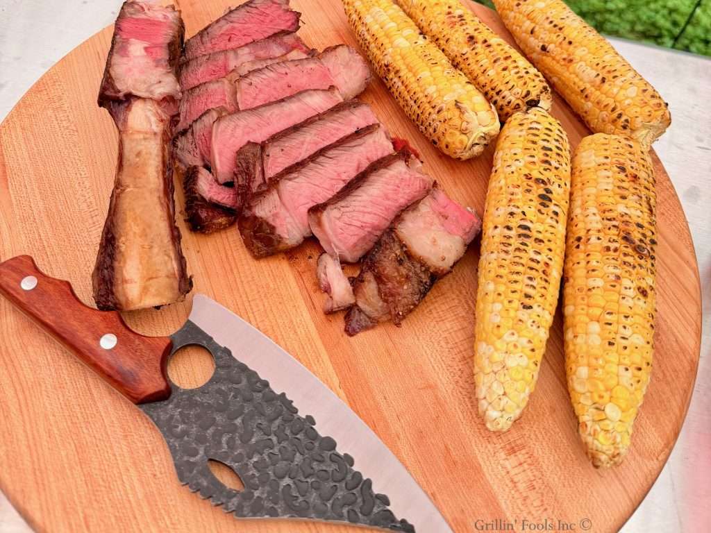 A carved bone in tomahawk steak cooked to medium rare, next to some grilled corn and a wicked looking knife on a blond cutting board