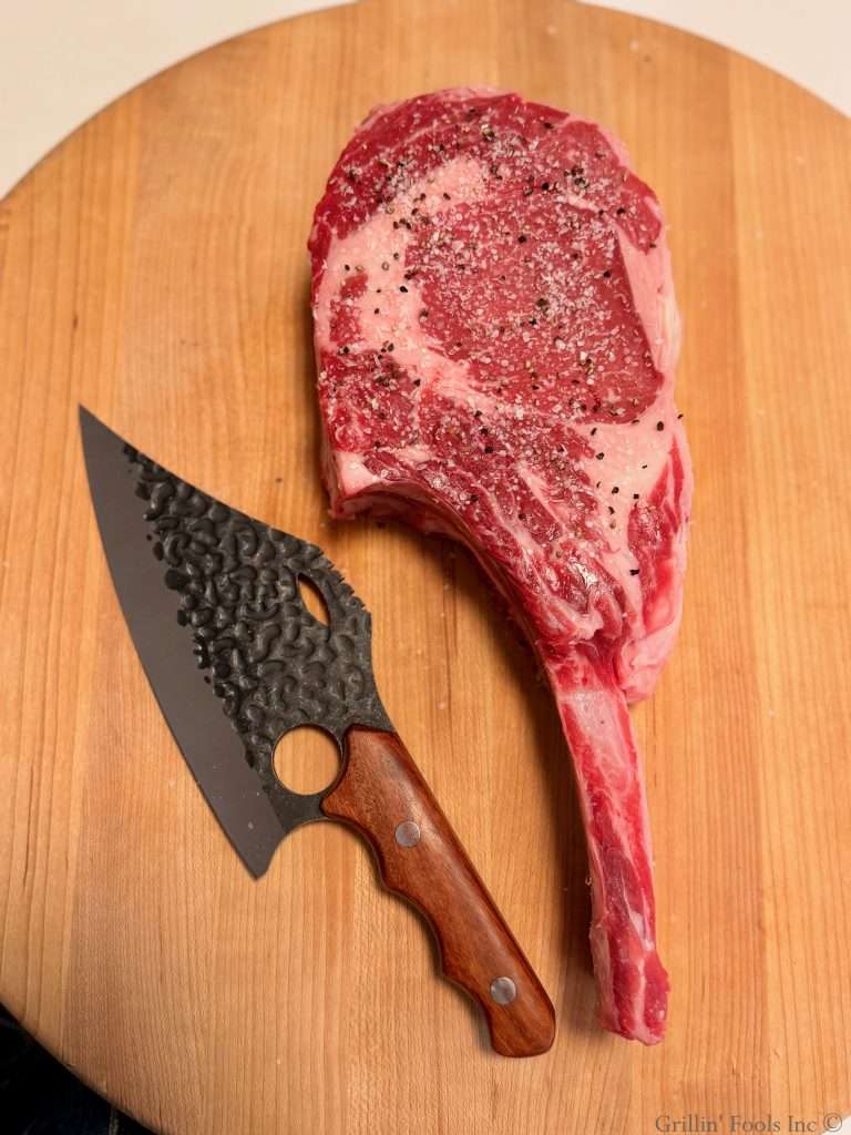 A raw tomahawk steak with a wicked looking knife on a blond cutting board