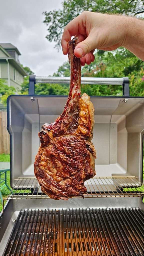 A hand lifts a long smoked and seared tomahawk steak from a barbecue grill outdoors, the bone exposed and meat hanging down.