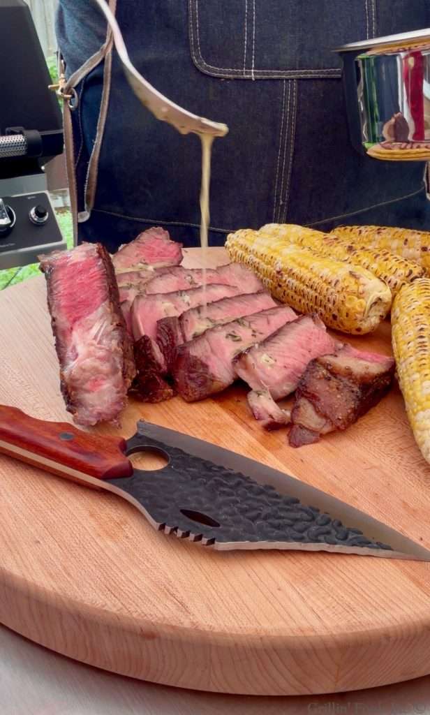 Drizzling cowboy butter over a sliced tomahawk steak next to some grilled corn on a cutting board