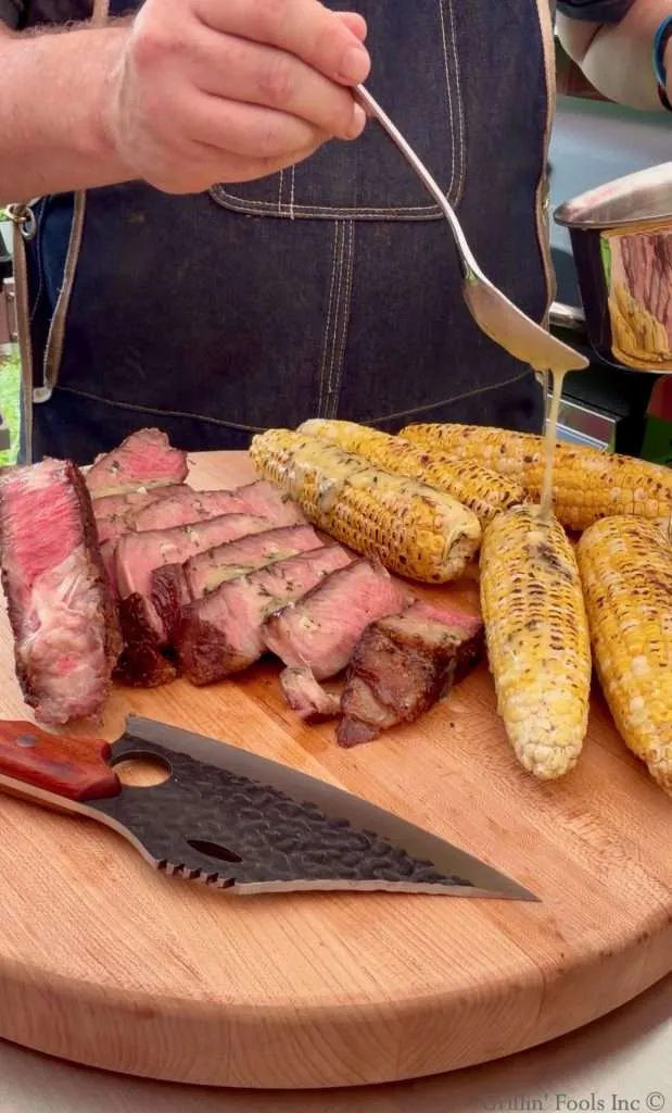 Drizzling cowboy butter over grilled corn next to a sliced tomahawk steak on a cutting board