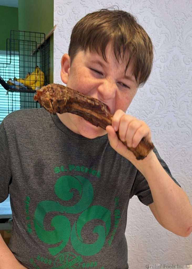 Boy wearing a gray shirt bites into a large rib bone at a white table in a home setting.