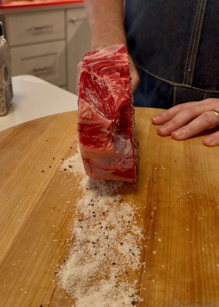 A thick slab of raw tomahawk steak with marbling stands upright on a wooden cutting board, with SPG seasoning scattered around and a person in a denim apron nearby