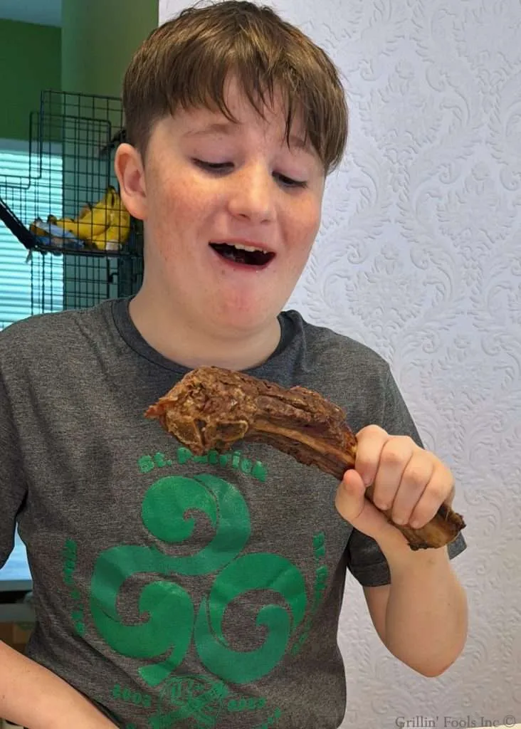Boy in a gray shirt holding a large chocolate brownie at a table, about to take a bite, with mouth open in anticipation.