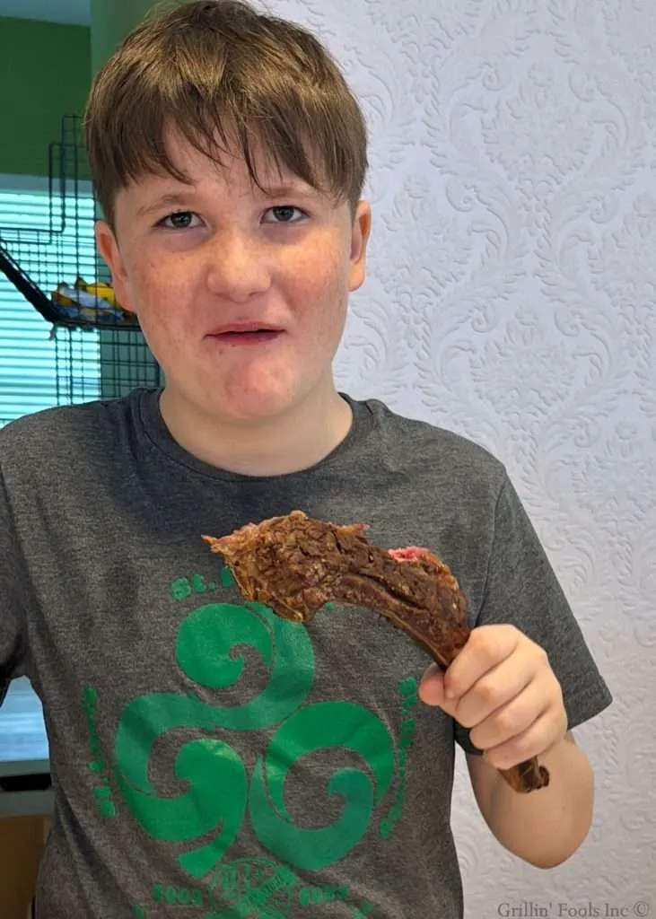 A young boy in a gray t-shirt holding a large chocolate-covered pastry near his mouth, indoors with a patterned white wall behind him.