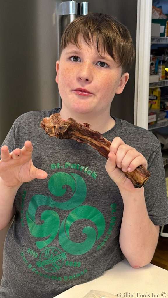Young boy with freckles holding a large rib bone, seated at a table in a kitchen and looking at the camera with a surprised expression.