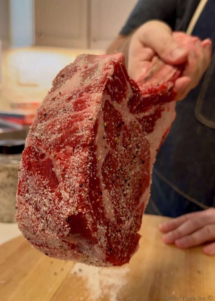 Salt-crusted large raw bone in ribeye held up in a kitchen, with the outer edge coated in salt, pepper and garlic seasoning.