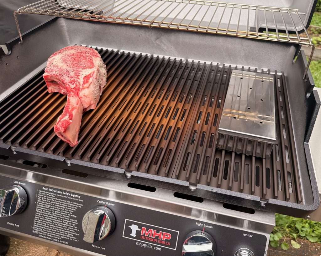 A seasoned, yet raw tomahawk steak on the left side of an MHP Hybrid grill with a metal smoker box on the right