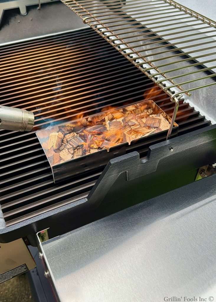 A fire box sitting full of pecan wood chips being ignited by a blow torch. The firebox is sitting on a gas grill