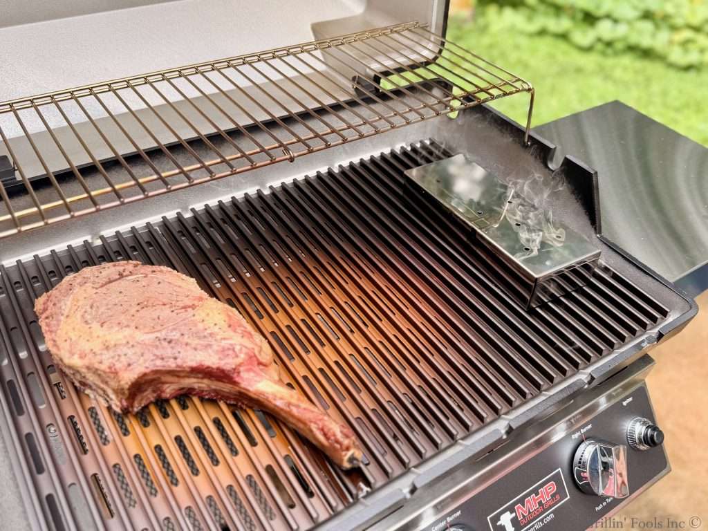 A tomahawk ribeye steak sits on the left side of a MHP Hybrid grill, with smoke rising from a smoker box on the right.