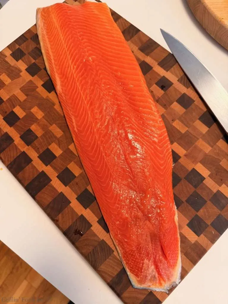 Raw side of salmon laid out on a checkered wooden cutting board with a knife nearby