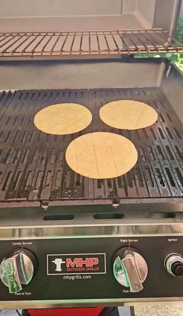 Three round tortillas cooking on a barbecue grill grate inside an outdoor grill.