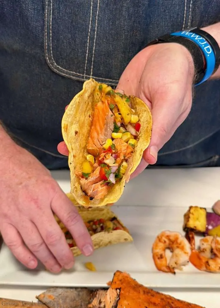 Close-up of a fish taco held in a hand, filled with grilled salmon, corn salsa, peppers, and cilantro in a warm corn tortilla.