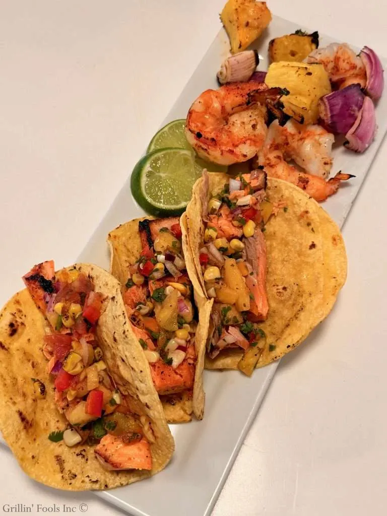 Salmon tacos with pineapple, corn salsa, onions, cilantro, on a white platter with lime slices.
