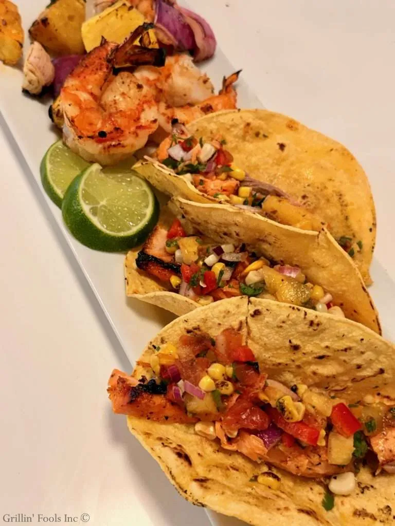 Grilled salmon tacos topped with corn salsa on a white platter, with lime wedges beside.