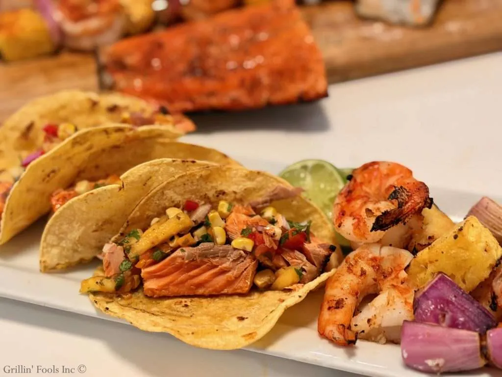 Salmon tacos topped with pico de gallo, served with lime wedges on a white plate.