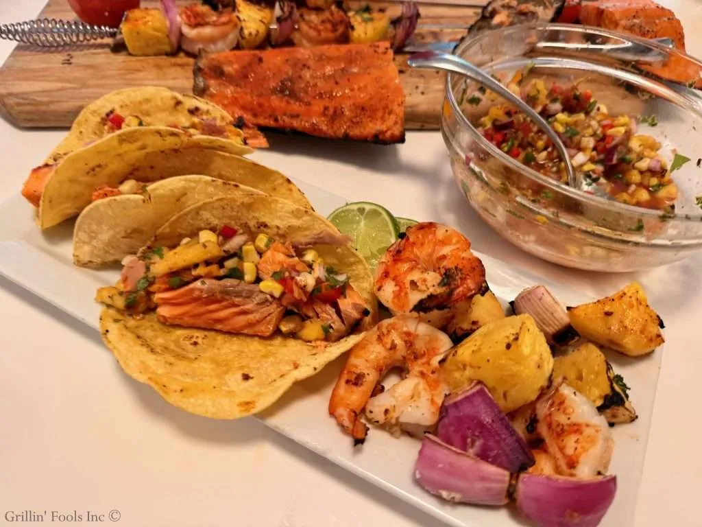 Plate of tacos filled with salmon, corn, peppers, and cilantro; lime wedges on the side, with shrimp and onions nearby for a Mexican-inspired platter.