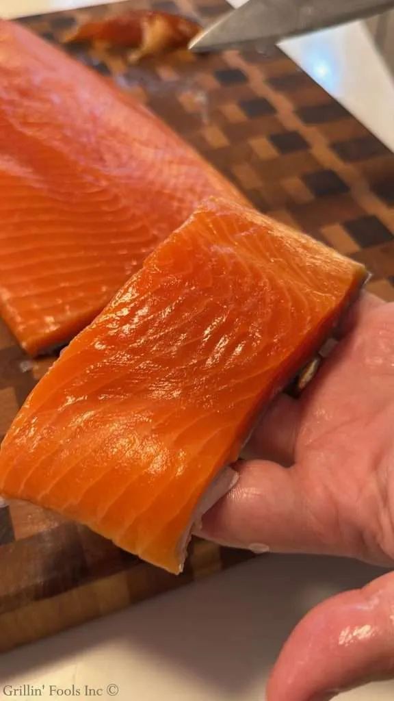 Close-up of a bright orange salmon fillet being sliced with a knife on a wooden cutting board surface.