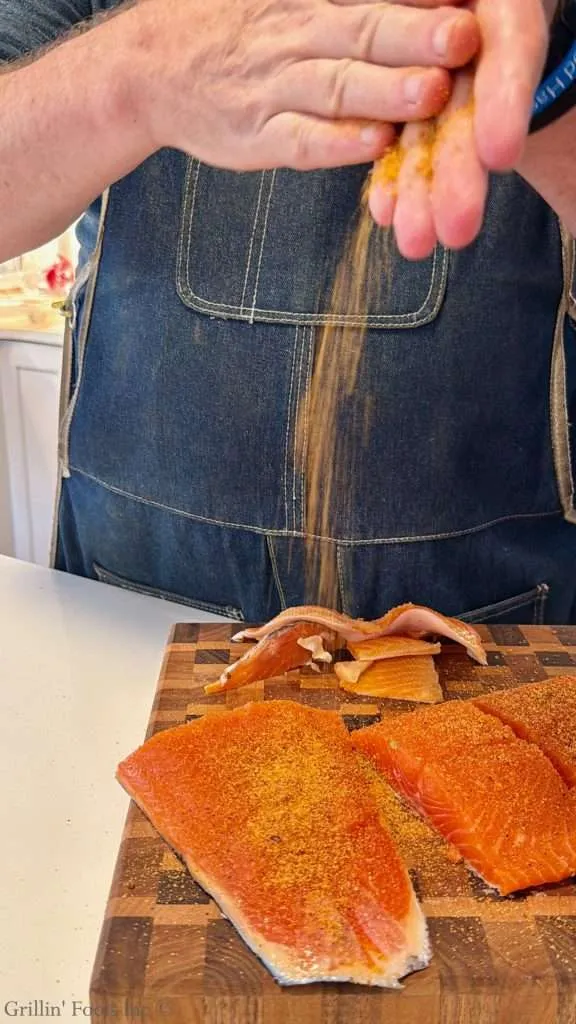 Hands in front of a denim apron sprinkling orange spice onto raw salmon fillets on a wooden cutting board adjacent to a cutting-board pattern.