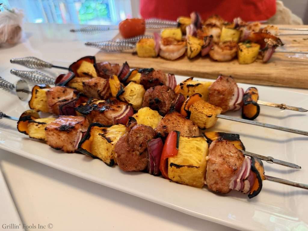 Skewers of grilled pineapple, red onion, bell pepper, and chunks of pork on a white platter with a wooden board in the background.