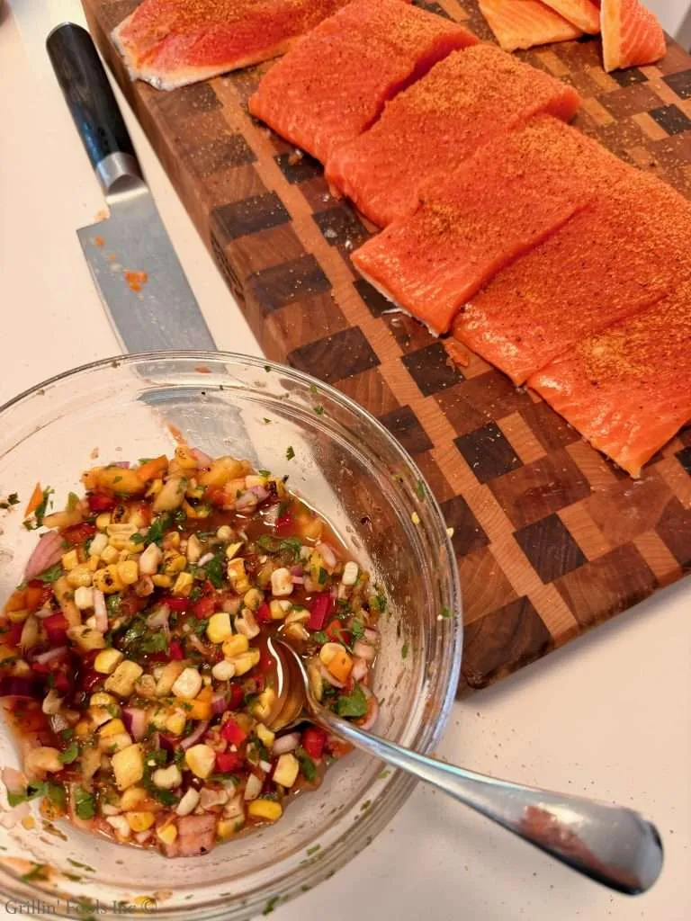 Salmon fillets seasoned with spices on a wooden cutting board next to a knife, with a bowl of chunky salsa nearby.