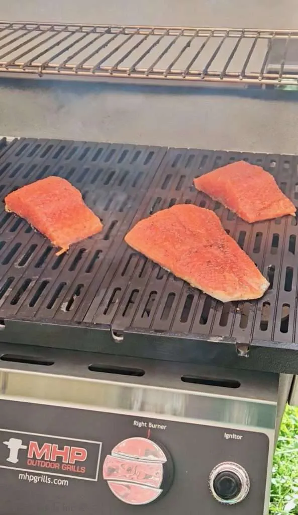 Three salmon fillets cooking on a black grill grate on an outdoor barbecue grill, seasoned with a pink crusted surface.