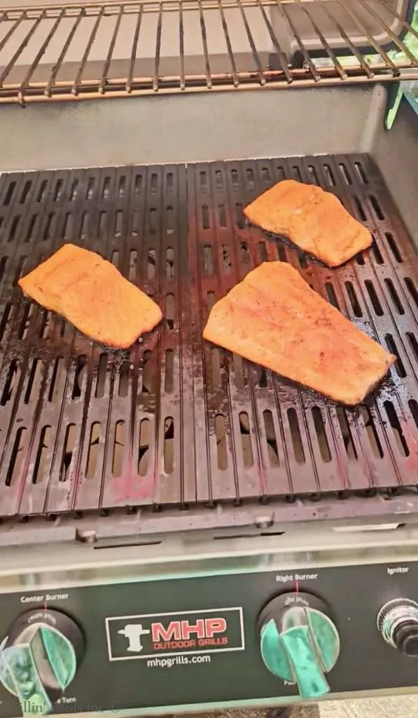 Three salmon fillets grilling on a metal BBQ grate over a barbecue grill.