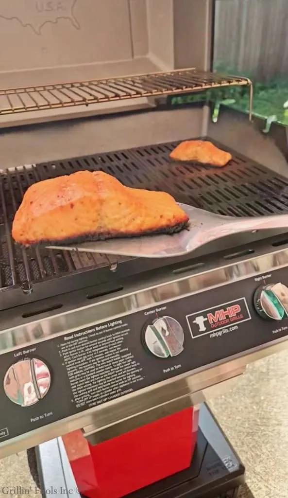 Two salmon fillets cooking on a gas grill, seasoned with orange seasoning on a ribbed grate.
