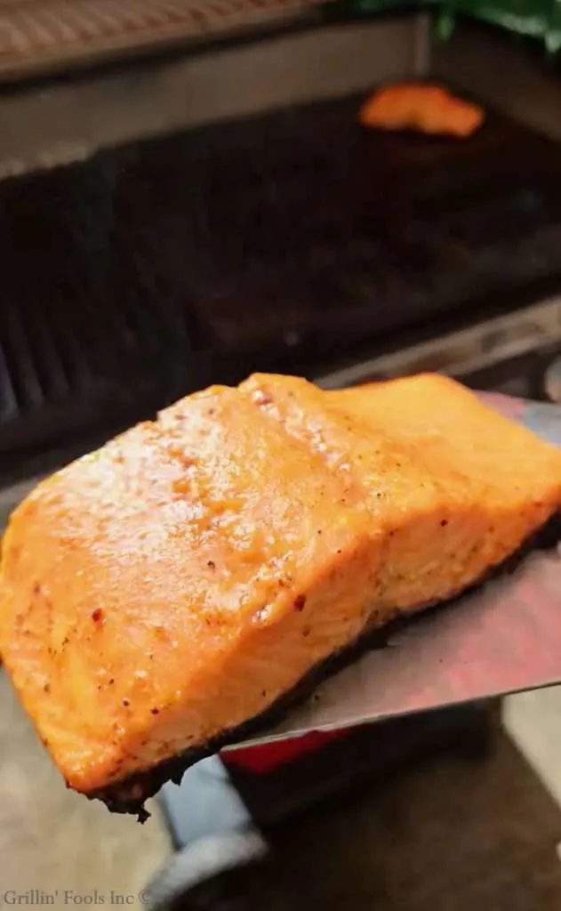 Close-up of a seasoned salmon fillet on a grill, orange-pink flesh with peppery seasoning and a crusty edge.