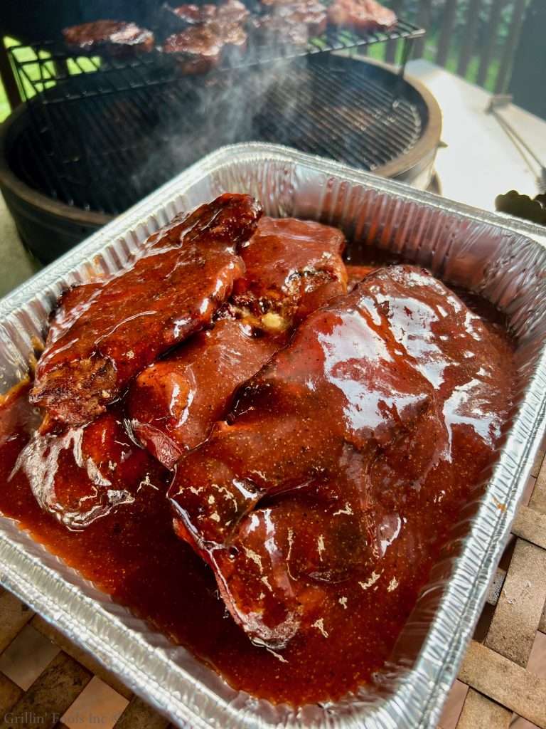 Barbecued pork steaks in a foil tray, glossy BBQ sauce with a grill visible in the background on a deck.
