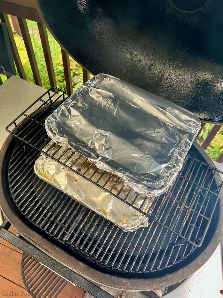 Two pans wrapped in aluminum foil sit on a grill grate with a dark smoker-style lid overhead, outdoors on a deck with greenery in view.