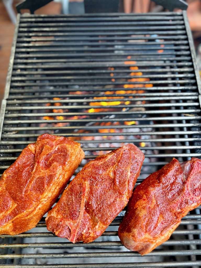 Seasoned pork steak grilling on a metal barbecue grate over hot coals with orange flames visible behind.