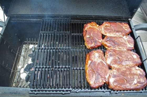 Outdoor barbecue grill with several pork steaks on the right side and charcoal briquettes on the left.