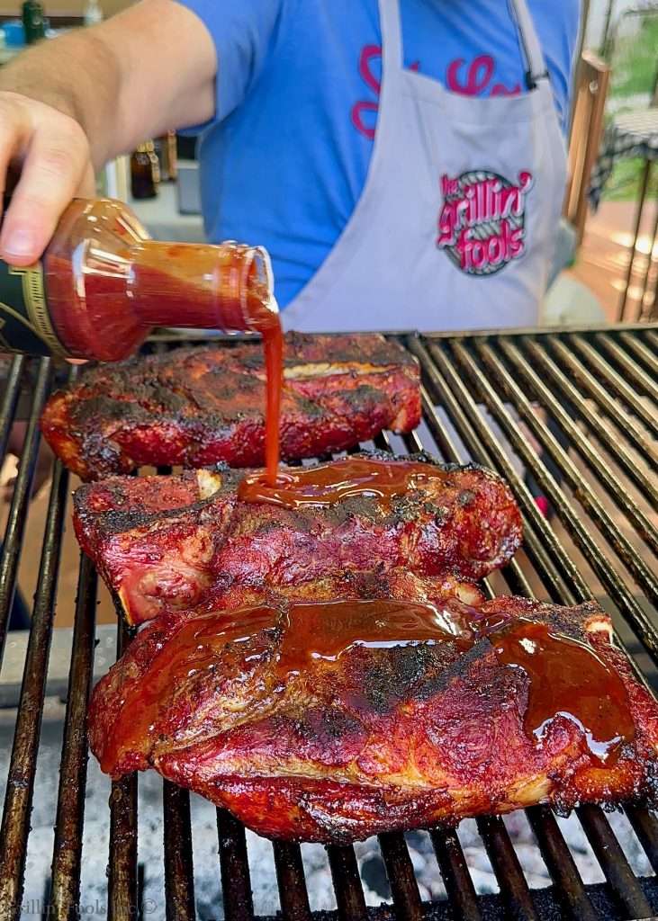 Barbecued pork steaks on a grill being glazed with dark barbecue sauce from a bottle.